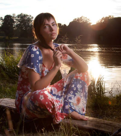 a young woman sitting on a log in front of a lake backlight from the sun lightened up with flashの写真素材