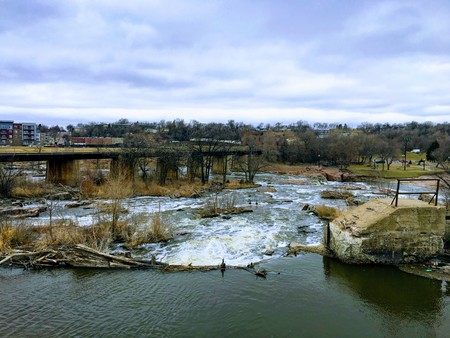 The Big Sioux River flows over rocks in Sioux Falls South Dakota with views of wildlife, ruins, park paths, train track bridge, trees and city in the surrounding area and backgroundの写真素材