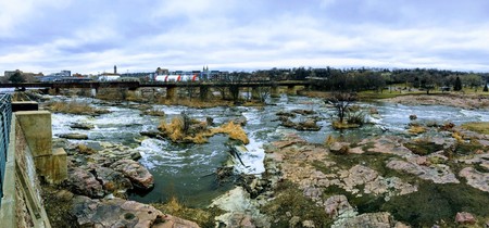 The Big Sioux River flows over rocks in Sioux Falls South Dakota with views of wildlife, ruins, park paths, train track bridge, trees and city in the surrounding area and backgroundの写真素材