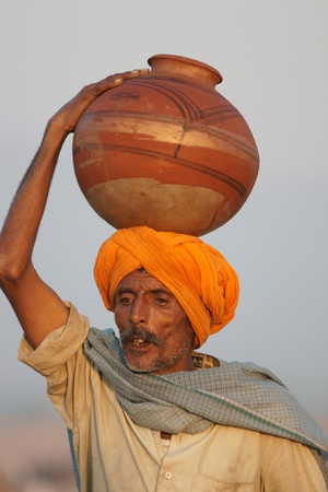 Pushkar, Rajasthan, India -  November 9, 2008: Indian man in orange turban carrying pot of water on his head at the Pushkar Fair, Rajasthan, Indiaのeditorial素材