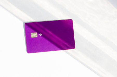 Plastic credit card with chip visible, on top of a table with soft lights and shadows. Purple card on white surface. Concept: finance, purchases, payments, loan, spending, investments and debts.の写真素材