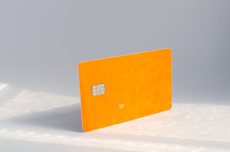 Plastic credit card with chip visible, on top of a table with soft lights and shadows. Orange color card on white surface. Concept: finance, purchases, payments, loan, spending, investments and debts.の写真素材