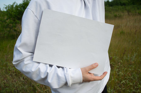 Woman in white sweatshirt holding mockup of white A4 sheet in hands, on background of green field.の写真素材