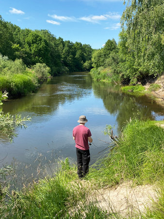 Man fishing by a calm river surrounded by lush green trees and grass under a clear blue sky, wearing...