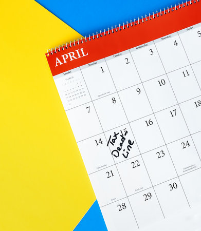 A close-up view of a calendar set to April, showing a tax deadline handwritten on the 15th day. The image includes a colorful yellow and blue backdrop.の写真素材