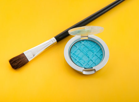 An eye-catching flat lay featuring a blue pressed eyeshadow compact paired with a makeup brush, all set against a bold, vibrant yellow background. Perfect for beauty and cosmetics themes.の写真素材
