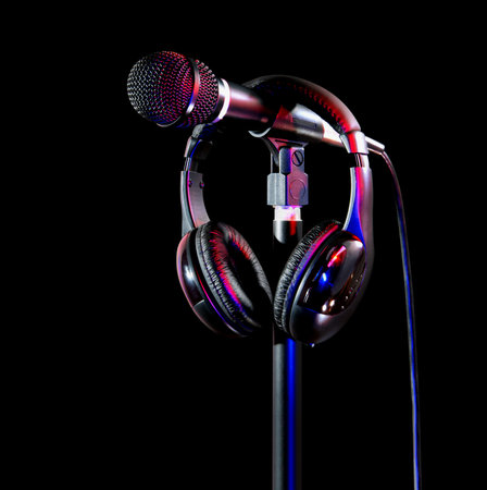 Studio equipment featuring a microphone and a pair of headphones resting on a mic stand. Captures a professional vibe ideal for musical, broadcasting, and recording themes in black and colorful lighting.の写真素材