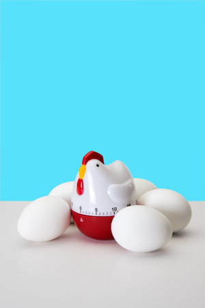 A chicken-shaped kitchen timer surrounded by white eggs sits on a clean white surface against a bright blue background. This playful still life concept showcases functionality and aesthetic appeal in kitchen decor.の写真素材