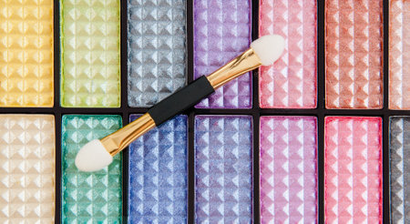 An up-close view of a vivid eyeshadow palette showcasing a range of bright, shimmery colors with a featured dual applicator brush laying on top. Ideal for cosmetics and beauty materials.の写真素材