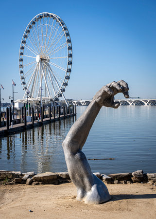 A sculpture of an arm protrudes from the sand in front of the Capital Wheel at National Harbor, Maryland with the Woodrow Wilson Bridge spanning the Potomac River in the background.のeditorial素材