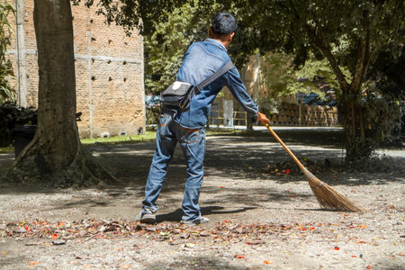 Man sweeps broom leaves in the garden in fall on ground. Colorful dry leaves: red, orange, yellow,...
