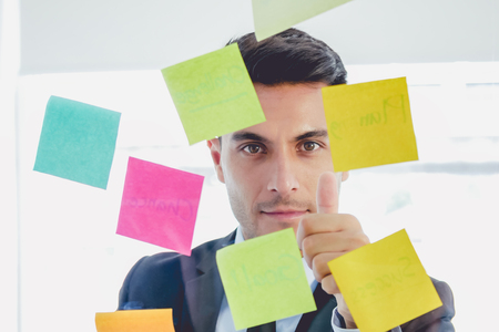 Handsome startup businessman looking straight sticky notes and thumb up in glasses stands in creative officeの写真素材