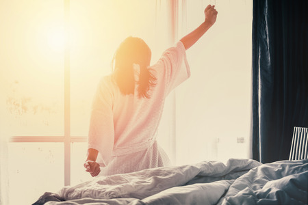 Woman stretching hands on bed after waking up while sitting on bed, entering a day happy and relaxed after good night sleepの写真素材