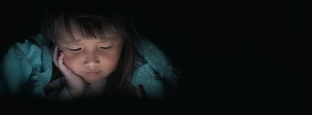 Little Girl watching tablets in bed at night with light flashes reflected from the screen,children using phones or playing tablet games and cartoon addiction smart phone concepts.wide banner.の写真素材