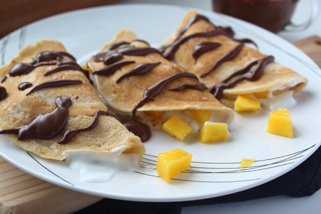 Healthy vegan wheat crepes decorated with chocolate. Wheat crepes with fresh mango and yogurt on plate and wooden background. Healthy breakfast. Horizontal image.の写真素材