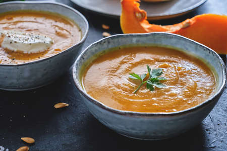 Homemade roasted squash cream soup with parsley leaf on the top on rustic bowl with raw squash pieces and seeds on black kitchen tableの写真素材