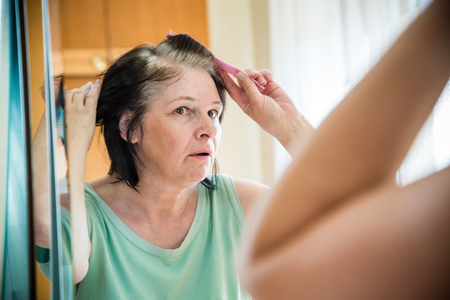 Senior woman checking her gray hair in the mirrorの写真素材