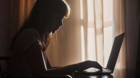 A woman typing and working on her computer with a beautiful warm light coming through her window.の素材