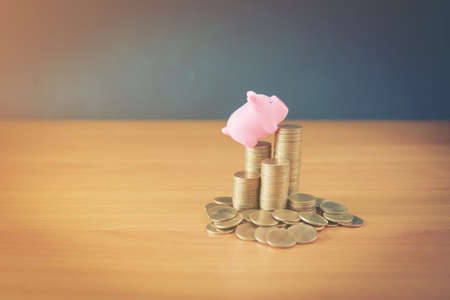 A piggy bank put on the stacking gold coins and blackboard house and clock on the vintage blue background, saving money for buy a new real estate or loan for planned investment in the future concept.の写真素材