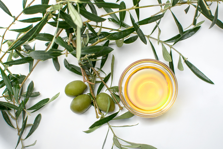 Glass bowl with olive oil, olives and branches with leafs. Close-up, top view with white backgroundの写真素材