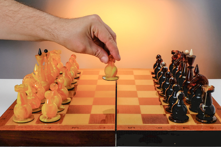 Chess game with amber chess figures on the board, first move. Hand of player holding white figure. With gradient gold background.の写真素材