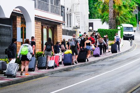 LLORET DE MAR, SPAIN - JULY 27, 2019: A group of tired tourists walks along the street with suitcases to the hotel on the Costa Brava. The concept of rest. Summer travel.のeditorial素材
