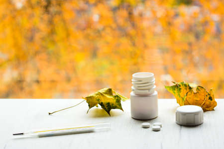 Thermometer, tablets from a white bottle and yellow leaves on white table, with autumn colors on background. Cold and flu season treatment concept.の写真素材