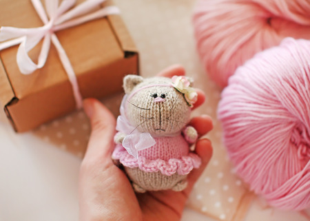 Handmade knitted toy. Knitted cat in a pink dress and bow in female hand on a background of gift box with ribbon and balls of yarn.の写真素材