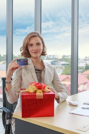 Smiling businesswoman showing credit card and red gift box in office - Christmas and Happy new year conceptの写真素材
