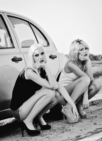 Two beautiful blonde girls sitting near their broken car and waiting for help  Black and whiteの写真素材