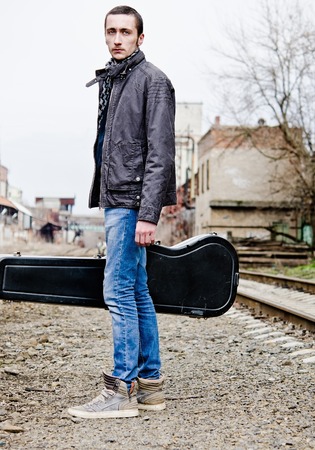Handsome young man with guitar case in hand standing amongst industrial ruinsの写真素材