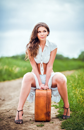 Beautiful young woman sits on a suitcase near rural roadの写真素材