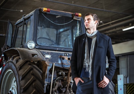 Fashion shot: a handsome young man wearing jeans and coat against the tractorの写真素材