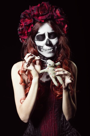 Young woman with calavera makeup (sugar skull) piercing a voodoo dollの写真素材