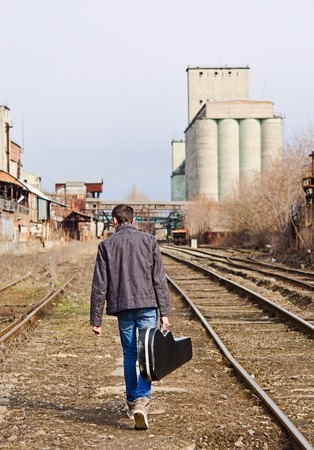 A young man with guitar case in hand going away by rails. Rear viewの写真素材