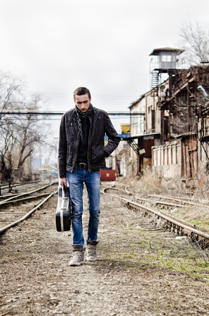 Sad lonely young man with guitar case in hand going by the railsの写真素材