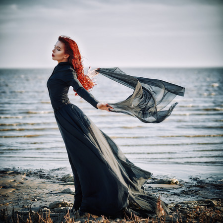 Beautiful sad goth girl with cloth in hands standing on the sea shoreの写真素材