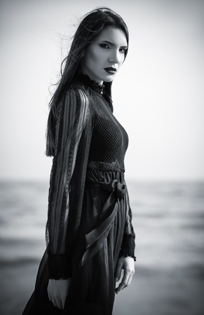 Beautiful sad gothic woman in black dress standing on the sea beach. Black and whiteの写真素材
