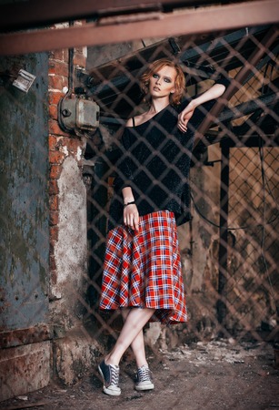 Portrait of a beautiful grunge girl in plaid skirt and sweater standing behind metallic latticeの写真素材