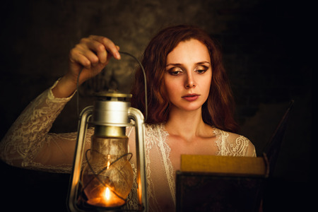 Beautiful red-haired young woman with kerosene lamp reading a bookの写真素材