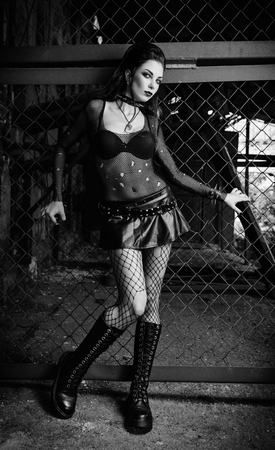 Portrait of the sexy goth (deathrock) girl dressed in holey blouse, skirt, bra and boots standing in industrial place. Black and whiteの写真素材