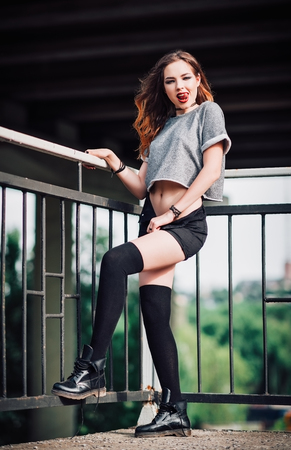 Outdoor shot: cheerful young rock girl in shorts, shirt and gaiters standing under the bridge and showing tongue の写真素材