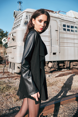 Fashion shot: portrait of the beautiful rock girl (informal model) dressed in black jacket and skirt standing at railroad (industrial area)の写真素材