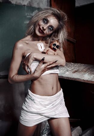 Strange mad girl with scary ugly doll in forsaken place (asylum). Crazy young woman (freak) dressed in messy bandages. Portrait of the smiling madman (schizophrenic)の写真素材