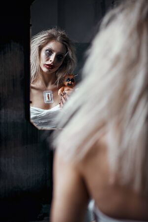Strange mad girl with horrible ugly doll in abandoned place (asylum). Crazy young woman (freak) looking into mirror. Portrait of the mental sick (madman)の写真素材