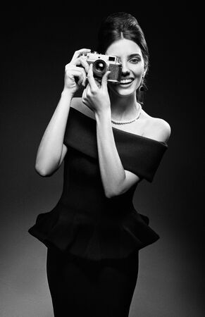 Retro shot: the beautiful young woman shooting by old film camera. Vintage portrait of smiling gorgeous girl in 60s style. Elegant lady in black dress and pearl necklace. Film grain effect, black and whiteの写真素材