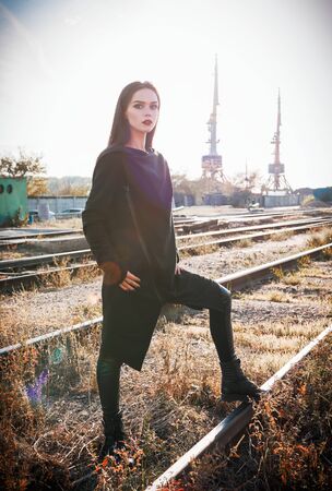 Fashion shot: portrait of the pretty rock girl (informal model) in tunic and leather pants standing at rails (industrial area)の写真素材