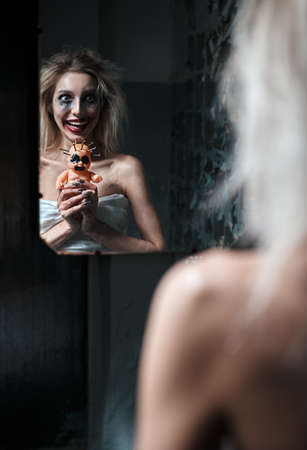 Weird insane girl with creepy ugly doll in abandoned place (madhouse). Crazy laughing young woman (freak) looking into mirror. Portrait of the mental sick (schizophrenic)の写真素材
