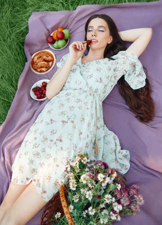 Picnic scene: lovely smiling girl lying on a plaid in park and eating strawberry. Outdoor portrait of beautiful young woman with closed eyes in dressの写真素材