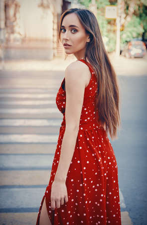 Beautiful girl in red dress stands at pedestrian crossing. Portrait of pretty young woman at the crosswalk. Rear viewの写真素材